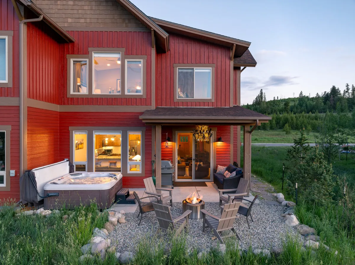 An inviting outdoor living space showcases a hot tub, fire pit, outdoor seating area, and large windows that connect the patio to the interior. The wooden siding and evening ambiance add warmth and a sense of togetherness.