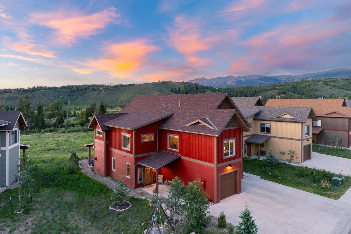 This image showcases a striking red house set amid lush greenery with a dramatic mountain backdrop and a vibrant, colorful sky. The setting evokes a sense of tranquility, natural beauty, and high-end mountain living, with ample space for outdoor gatherings and relaxation.