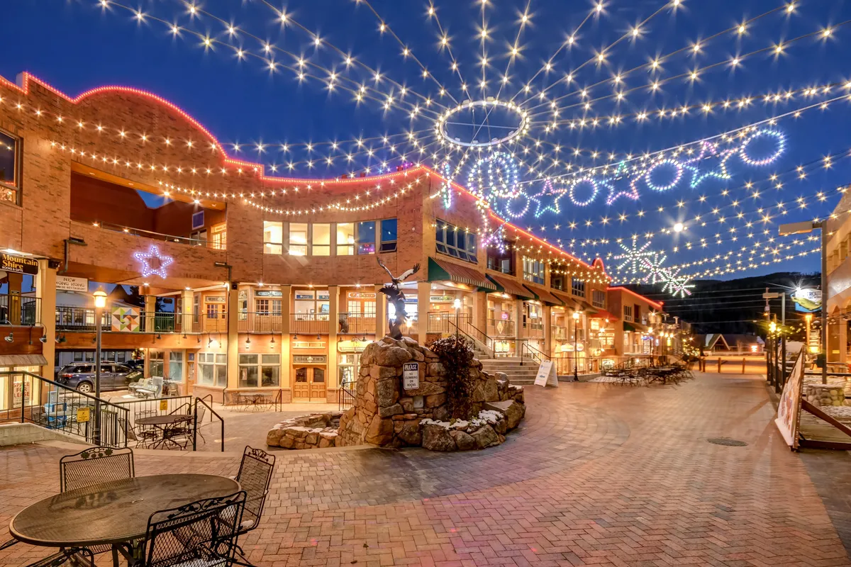 An inviting outdoor plaza is illuminated by overhead string lights and decorated with festive star motifs, with a central stone fountain creating a focal point. The scene suggests community events, evening gatherings, and the celebratory nature of the area.