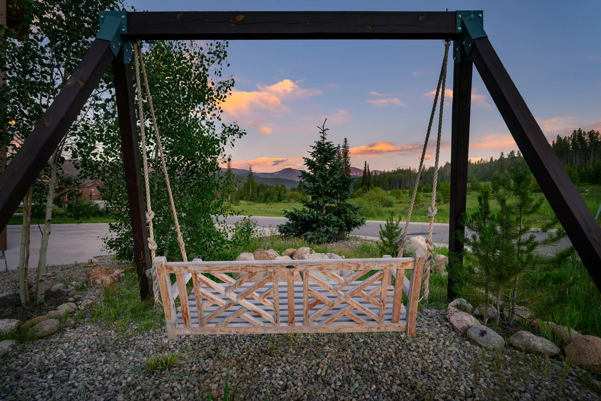 A serene outdoor setting features a wooden swing surrounded by lush greenery, with mountains providing a breathtaking backdrop. The space feels tranquil yet social, inviting relaxation or conversation with friends.