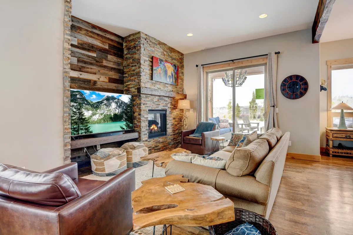 This image captures a warm, inviting living room featuring a striking stone fireplace, large window with a scenic view, a TV screen, and comfortable seating arranged for conversation. Artwork and a wooden coffee table add a touch of mountain-chic style.
