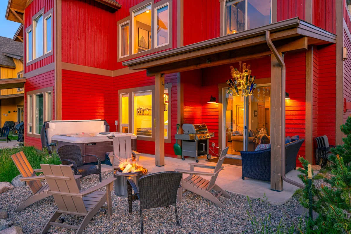 An inviting outdoor patio area with the red exterior of the house, a BBQ gas grill, a fire pit (Solo Stove), a hot tub, and comfortable outdoor seating. The space is arranged for socializing and enjoying meals outside with mountain views.
