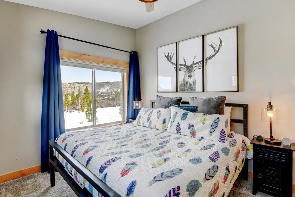 A guest bedroom with a bed adorned in a colorful quilt, deer artwork above, and a large window providing a bright, airy feel and beautiful mountain scenery.