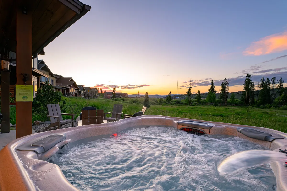 An exterior scene focusing on a steaming hot tub set on a patio, surrounded by lawn chairs and an expansive mountain backdrop at sunset. The tranquil atmosphere invites relaxation and stargazing, echoing a memorable evening soak.