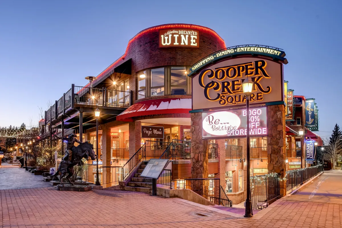This exterior scene captures the lively Cooper Creek Square, featuring visible signage for shops, a wine shop, and an outdoor seating area. The area is bustling, with elements that suggest shopping, dining, and socializing opportunities in a festive atmosphere.