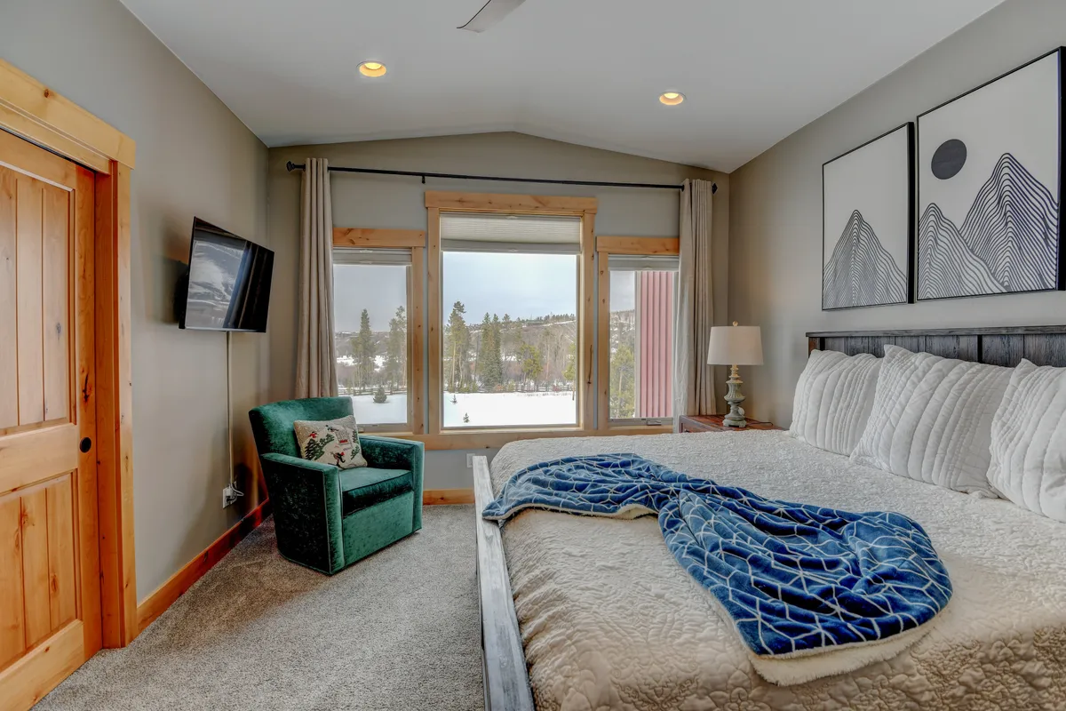 A spacious bedroom featuring a large bed with textured bedding, mountain artwork on the wall, a window offering a view of the outdoors, and a green accent chair positioned for reading or relaxing by the window.