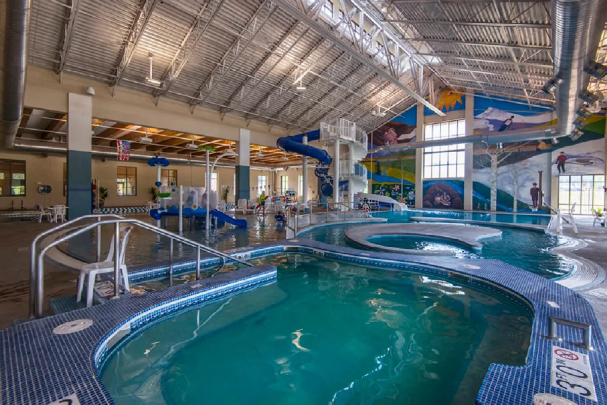 A lively indoor recreation center featuring a large swimming pool with water slides, a colorful mural, and families enjoying time together. The space is vibrant and energetic, perfect for year-round recreation and wellness.