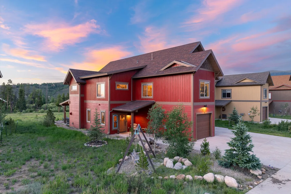 The image showcases the striking red exterior of a spacious mountain chalet nestled among lush greenery in Fraser, Colorado. A vibrant play structure sits in the yard, inviting children to play while adults relax on the porch and take in the sunset views. The setting conveys a sense of warmth, community, and family fun, perfect for gatherings and outdoor activities.