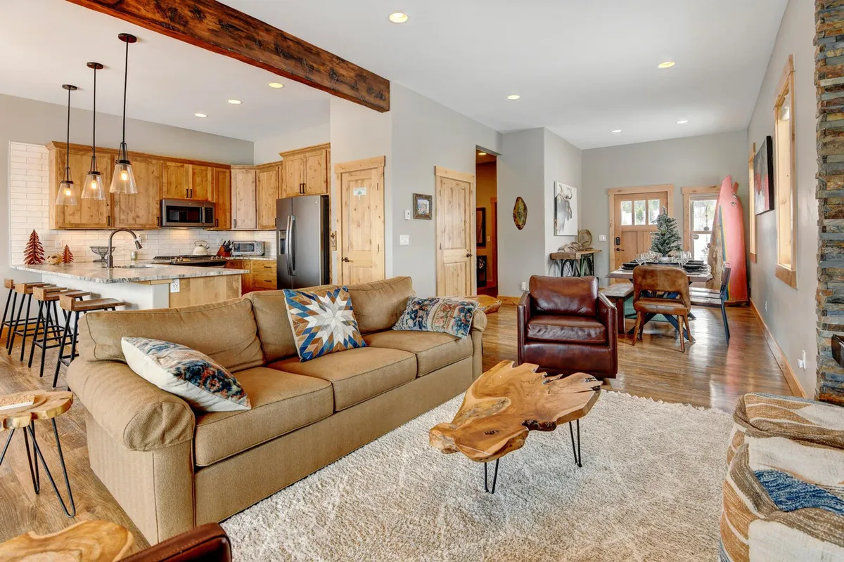 A wide-angle view of an open-concept interior where a comfortable sofa, rustic wood accents, a kitchen island, and a dining area are seamlessly connected. Art pieces and abundant light create a welcoming, cohesive social space.