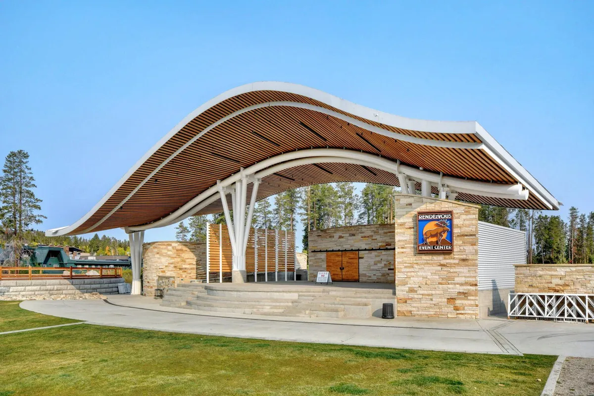 An exterior shot of a curved roof structure with a stone facade and wooden details, likely representing Hideaway Park. The space suggests community gatherings, outdoor concerts, and festive events set against a mountain backdrop.